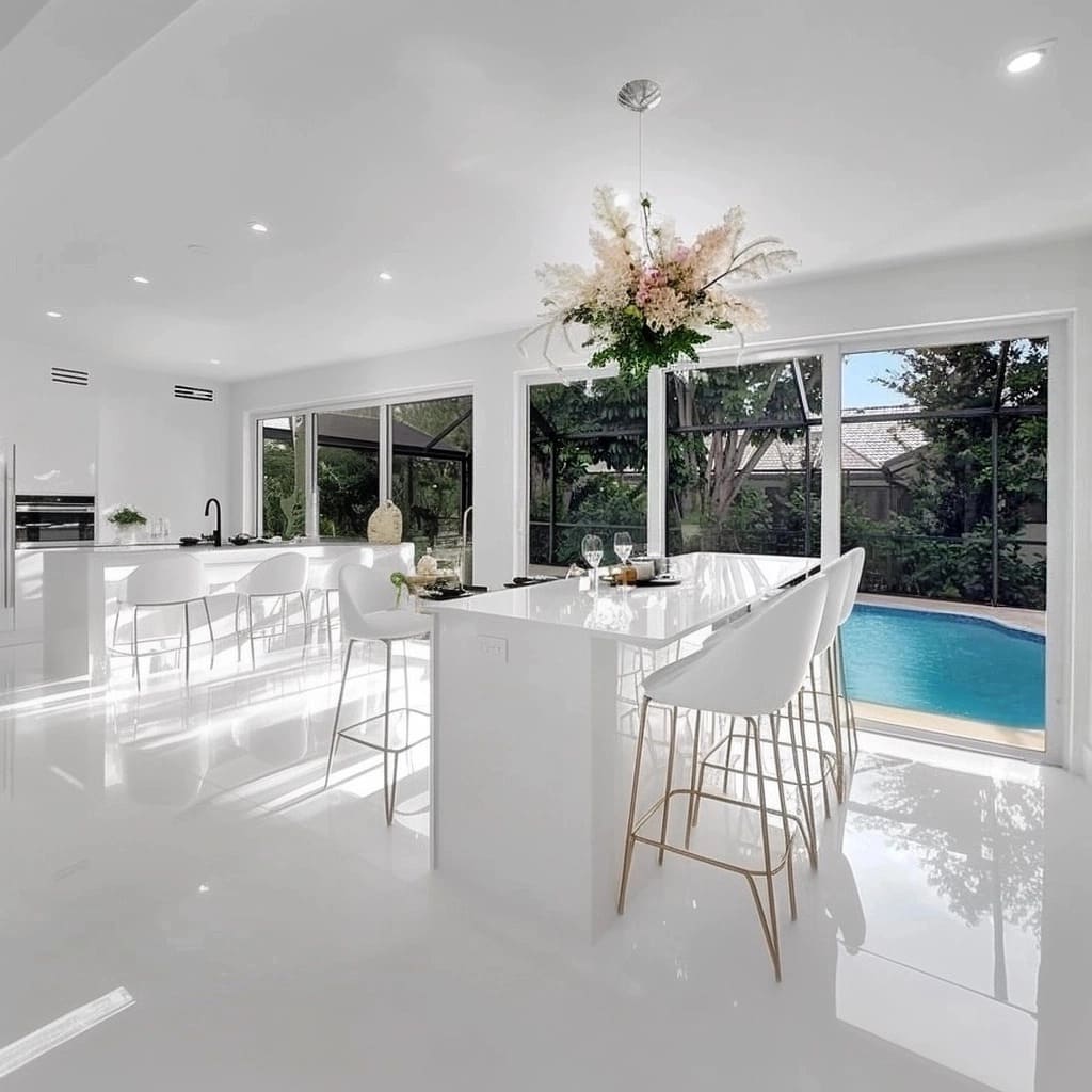 A long island in glossy white finish takes center stage, accompanied by sleek white bar stools with thin, gold-colored legs that introduce a subtle touch of luxury.jpg