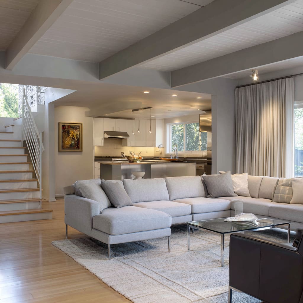 Quiet front room design under exposed beams, linen sofa with chrome legs, glass coffee table, and satin kitchen accents